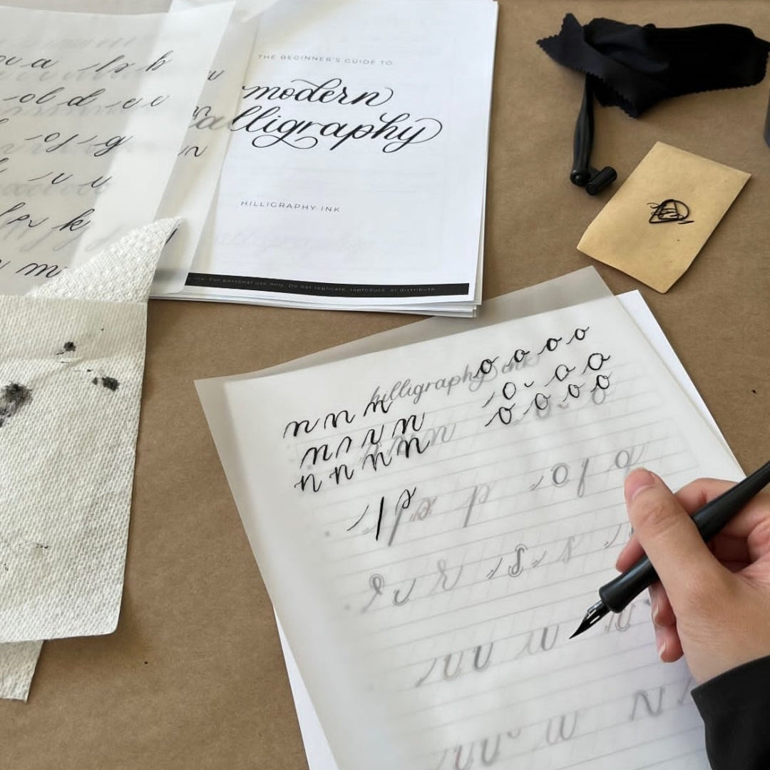 Person practicing calligraphy with a pen on a piece of paper, surrounded by writing materials on a table.