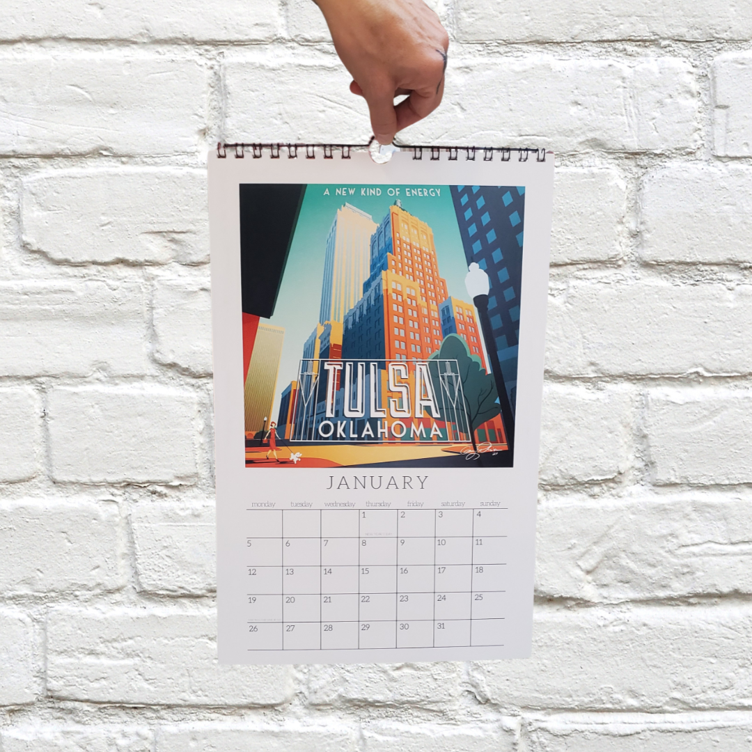 Hand holding a calendar with a 'Tulsa, Oklahoma' design against a white brick wall.