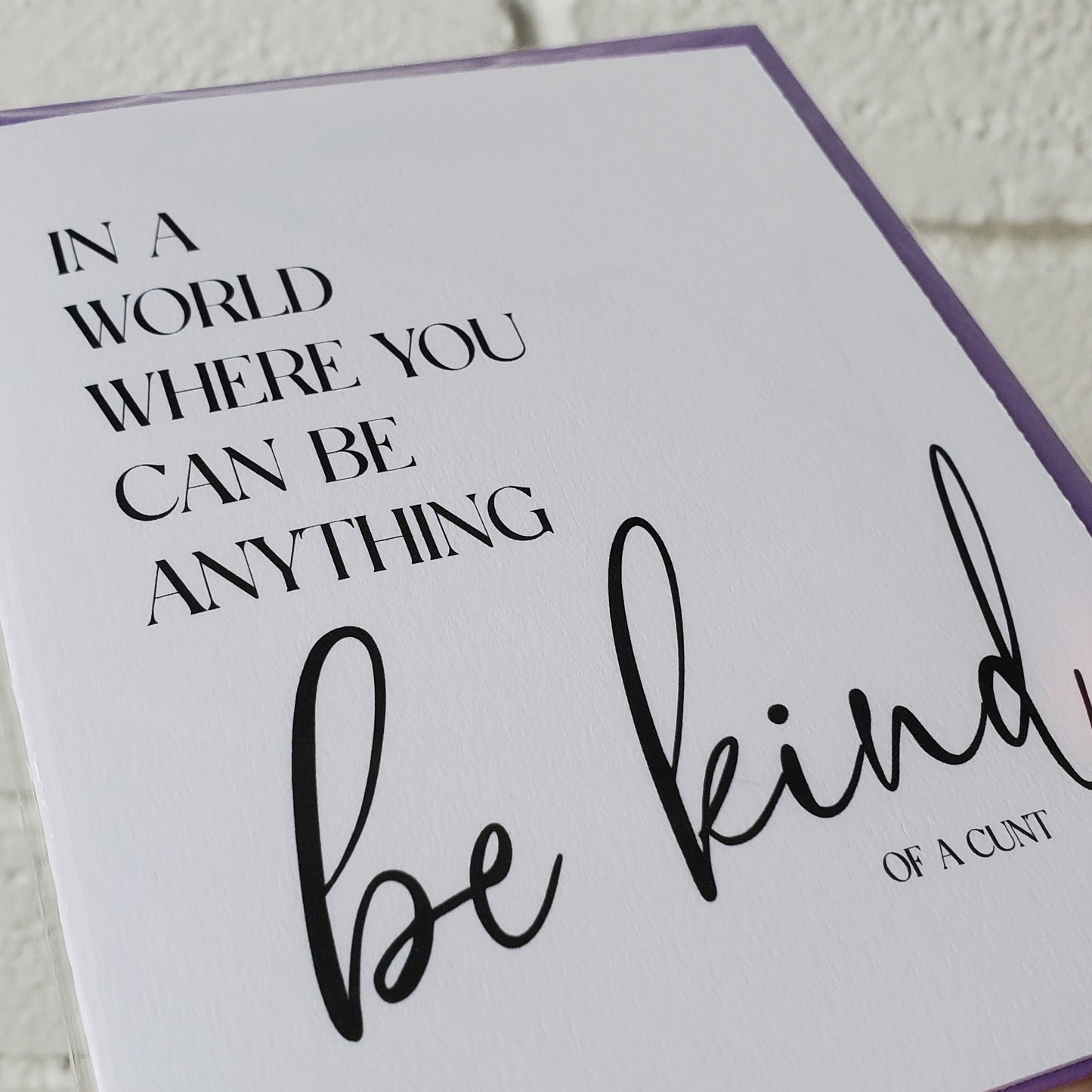 Card with text 'In a world where you can be anything, be kind of a cunt' held against a textured wall.