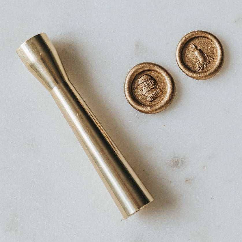 A brass wax seal stamp with a mid-century modern design, featuring a knitted mitten and an Advent candle with a simple wreath, placed on a white surface.