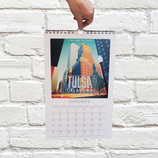 Hand holding a calendar with a 'Tulsa, Oklahoma' design against a white brick wall.