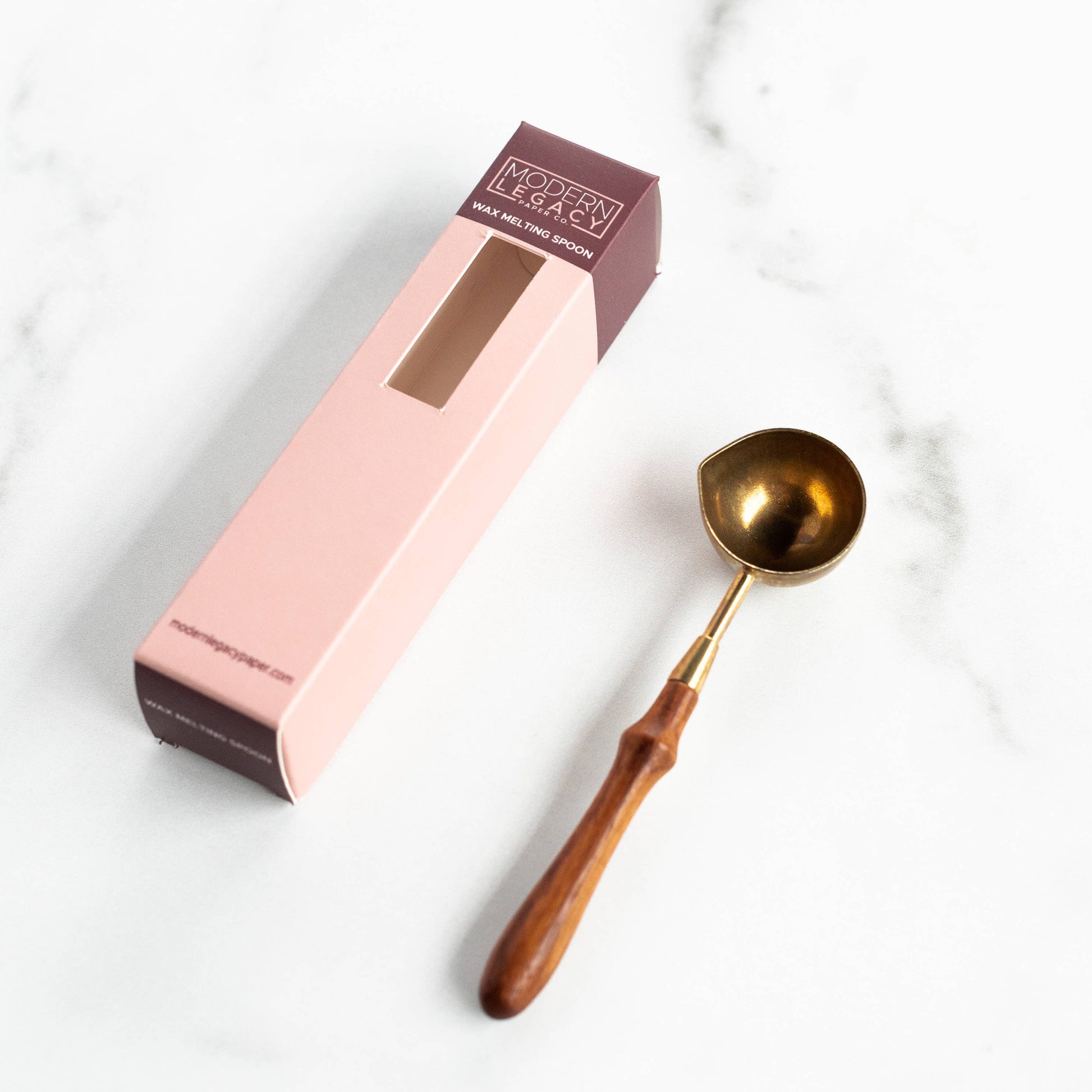 A brass spoon with a rosewood handle alongside its pink packaging box, placed on a marble surface.